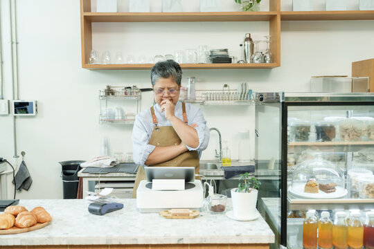 Asian senior female cafe owner at counter with hand on chin looking at tablet reflecting on small business decisions confidently managing independent post-retirement coffee shop.