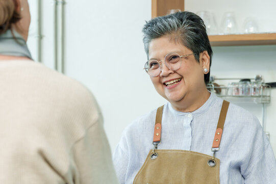 Asian senior female barista adult laughing joyfully while talking with customer at cafe counter representing positive customer interaction in cozy local family coffee shop, Concept small business