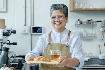 Cheerful elderly Asian woman barista holding tray with croissant and tea, smiling in cafe, expressing joy, pride, hospitality, enthusiasm, reflecting charm of small coffee shop, Concept small business