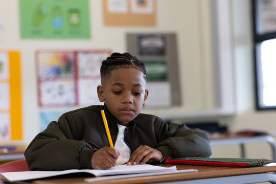African American boy sitting in classroom writing in notebook with pencil and pencil case