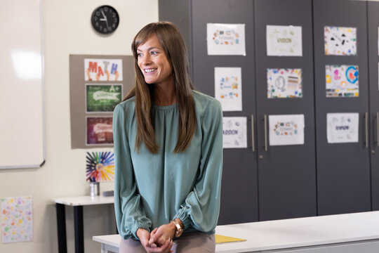 Woman leaning at desk in classroom smiling toward right with whiteboard and color pencils