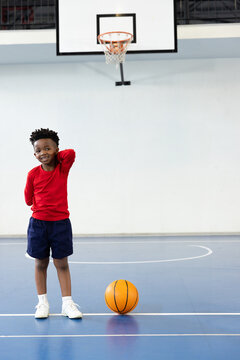African American boy standing on blue court in gymnasium under hoop next to basketball, copy space