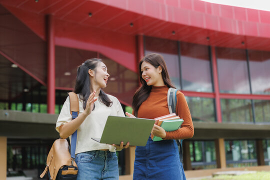 Two cheerful university students talking and walking outdoors with laptop and books