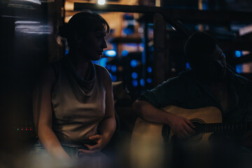 A woman listens attentively to a musician playing acoustic guitar in a dimly lit setting, creating...