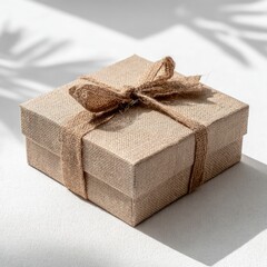 Square, light-brown gift box with burlap ribbon, casting shadow on bright surface