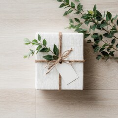 A simply wrapped white gift box adorned with twine, a blank tag, and sprigs of greenery rests on a light wood surface