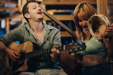 Young friends gather for a live music session, with a guitarist and tambourine player, creating a lively atmosphere in a cozy wooden setting.