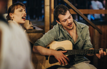 A young man strums an acoustic guitar as a woman sings enthusiastically beside him in a warmly lit, wooden-furnished cafe, capturing the essence of live music and creativity.