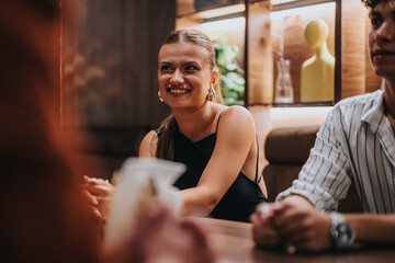 A cheerful woman smiles while seated at a table in a cozy restaurant, engaging in a lively social gathering with friends.