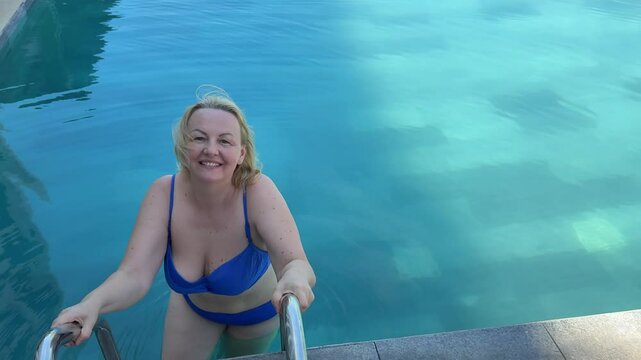 adult plump woman laughing enters the pool to relax swim blue swimsuit blond hair beautiful smile 45-55 years old