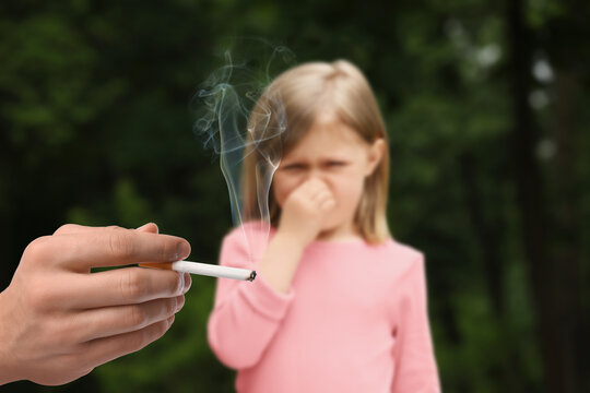 Passive smoking. Little girl covering her nose with hand because of man smoking nearby outdoors. Selective focus