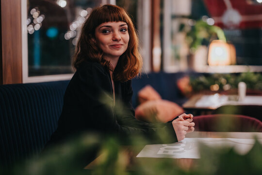 A woman with a warm smile sits in a cozy cafe, surrounded by soft lighting and lush plants, creating a relaxed and inviting atmosphere.