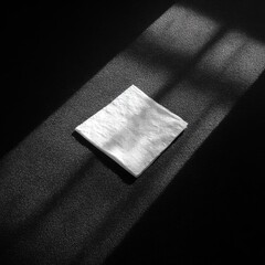 A pristine white cloth rests on a dark surface, illuminated by a narrow sunbeam slicing through a shadowed room, creating strong contrasts and highlighting its texture