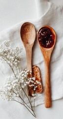Two wooden spoons, one holding dark red preserve, rest on linen with small white flowers and a piece of biscuit