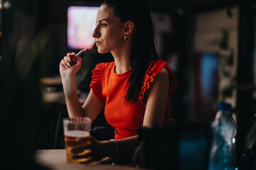 Young woman in red top seated in a dimly lit restaurant, holding a drink and contemplating,...