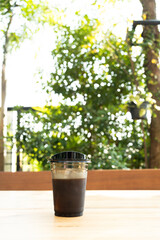 A plastic cup with iced black coffee sits on a table in a bright from a nature with a blurred green nature background.