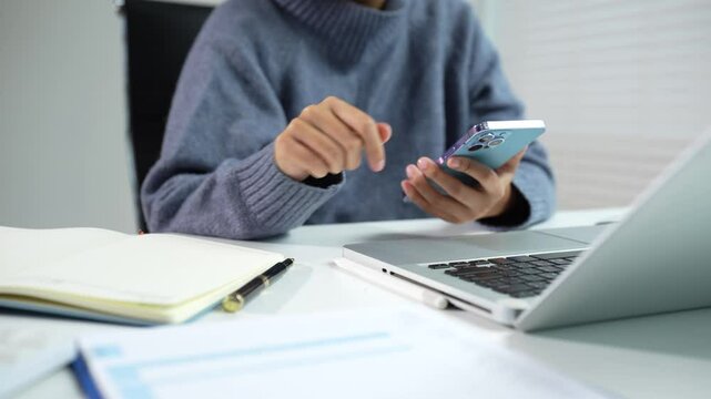 Employee, taking notes sitting at desk using smartphone and laptop searching for information at work. - Powered by Adobe
