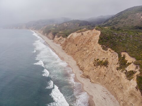 Aerial view of a rugged coastline  of Point Reyes National Seashore, Bolinas, California, USA. Waves crash on the sandy beach below steep cliffs, with fog blanketing the hills in the background.