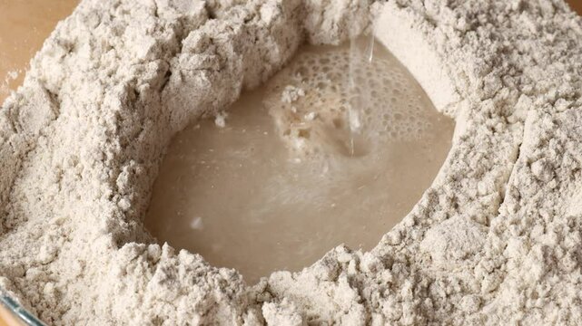 pouring water into flour in deep well for preparing dough starter to bake homemade bread showing closeup of mixing process concept of bakery shop cooking blog traditional baking