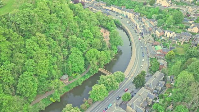 Real-time drone slowly descends above the curving River Derwent and riverside road in Matlock Bath, revealing cars, footbridges, and dense greenery along this iconic Derbyshire landscape.