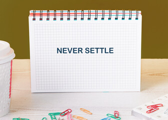 Concept words NEVER SETTLE in an open notebook surrounded by colorful paper clips and a coffee cup