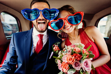 Happy couple in oversized heart sunglasses pose playfully inside a vintage car with a bouquet, celebrating a wedding or special event with joy and humor.