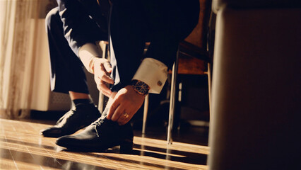 Man in a suit tying his black dress shoes, showing hands, watch, ring, and cuff. Preparing for a formal event, business meeting, or wedding day.