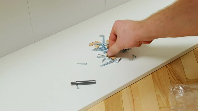 Carpenter hand picks up wooden dowel pins from screws pile on plywood panel closeup. Man takes fastening for furniture assembling working on floor at home