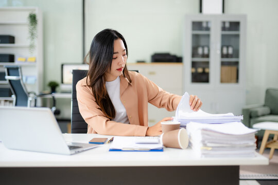 Asian businesswoman focused on sorting stacks of paperwork at her desk with a laptop and coffee cup.