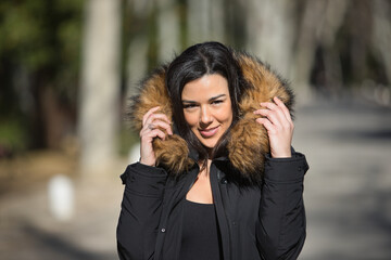 Portrait of a young, beautiful, dark-haired woman, looking at the camera, wrapping herself up with the fur collar of her coat, on a cold and sunny day. Concept winter, cold, autumn, weather.