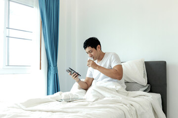 Asian man using smartphone while drinking almond milk in bed after waking up in morning in bedroom....
