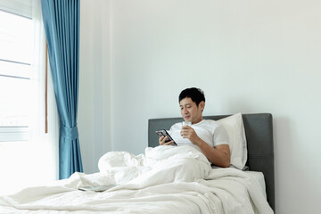 Asian man using smartphone while drinking almond milk in bed after waking up in morning in bedroom....