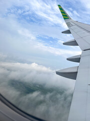 Airplane wing soars above fluffy clouds with patches of green landscape visible below, set against a bright blue sky in daylight, viewed from the window seat.