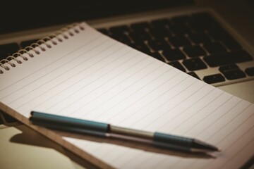 Flat design notebook with ballpoint pen is resting on laptop keyboard over desk with warm lighting