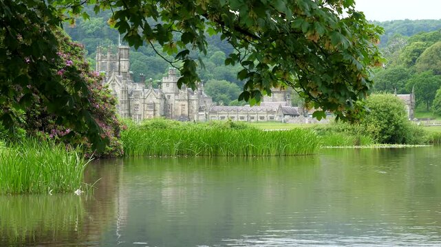 Part of Margam mansion view through tree branches in the park in South Wales, summer greenery and blossom around pond or lake. Local council property.