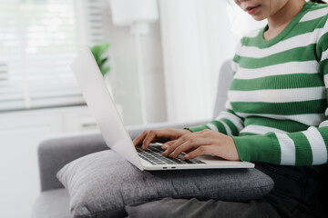 Naklejka premium A young woman wearing headphones and glasses is sitting comfortably on a sofa. She looks at her smart phone and laptop.
