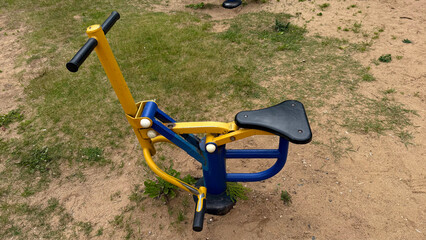 Colorful outdoor exercise equipment for children, people on a sandy playground, blue and yellow metal fitness machine with seat and handlebars
