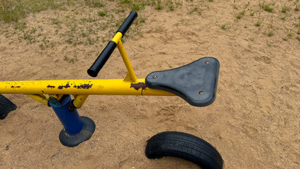 Old yellow metal seesaw with black handles and worn paint on a sandy playground, vintage outdoor children’s equipment
