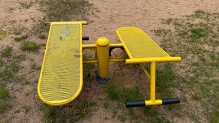 Outdoor yellow metal fitness bench for leg and abdominal workouts in a sandy playground with grass patches
