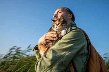 Man traveler strolling down with tabby cat in collar on sunny day on beach outdoors under blue sky....