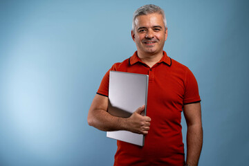 Smiling mature businessman holding closed laptop against blue background