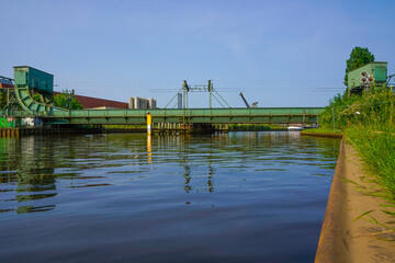 An industrial bridge over a tranquil canal with a clear blue sky and green banks. The bascule...