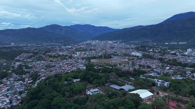 Aerial footage of Ibague at dusk, showcasing sprawling cityscape and lush greenery under soft twilight. Mountains form a majestic backdrop. The atmosphere is serene. Soccer stadium at distance.
