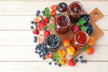 Tasty sweet jams in jars surrounded by ingredients on light wooden table, flat lay. Space for text