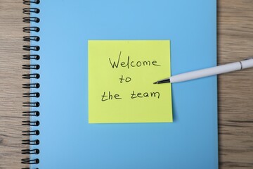 Sticky note with phrase Welcome To The Team, pen and notebook on wooden table, top view