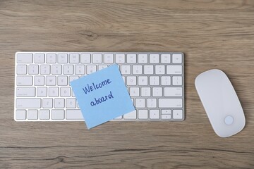 Sticky note with phrase Welcome Aboard, computer keyboard and mouse on wooden table, flat lay