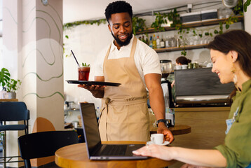 Smiling African American waiter serving coffee and juice to female customer sitting in cafe.