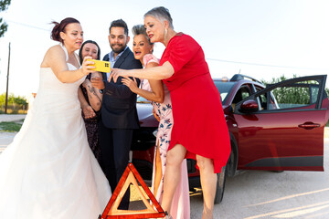 Wedding guests taking selfie with bride and groom next to broken car