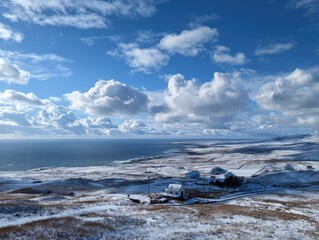 Snowy coastal landscape with a cluster of houses under a vibrant blue sky dotted with fluffy white clouds