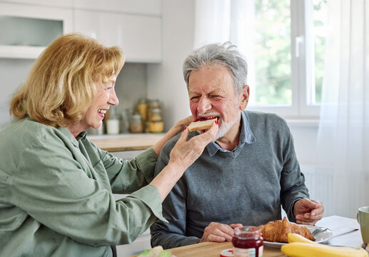 Portrait of an elderly senior couple having breakfast sharing a donut dessert at home. Happy healthy affectionate senior couple eating and sitting at kitchen table having fun enjoying morning meal tog